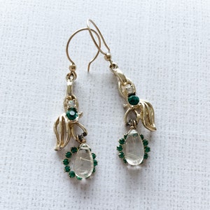 May include: A pair of gold-tone drop earrings featuring a curved metal design accented with small clear and emerald green rhinestones. Each earring suspends a clear, teardrop-shaped stone surrounded by a halo of tiny emerald green rhinestones.