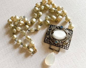 May include: A necklace featuring a strand of cream-colored faux pearls connected to a rectangular, antiqued silver-tone metal pendant. The pendant contains a central white oval cabochon, two small blue accent stones, and a dangling teardrop-shaped bead.