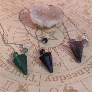 May include: Three crystal pendulums with silver chains rest on a wooden surface with astrological symbols. One pendulum is green, one is black, and the other is purple. A large, light-colored crystal sits above the pendulums.