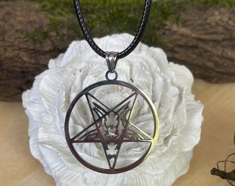 May include: A silver-colored pendant necklace featuring a pentagram design within a circle, suspended from a black cord. The pendant has intricate details and is displayed against a white, textured background.