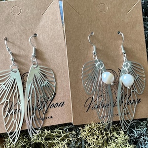 May include: Two pairs of silver butterfly earrings with a pearl accent. The earrings are displayed on a brown background with the text "Vampire Moon" and "Mystic in the Air".