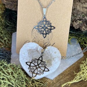 May include: Two silver-colored Celtic knot necklaces. One necklace is displayed on a brown card with a drawing of a woman. The other necklace is placed on a white heart-shaped geode. The background includes moss and wood.