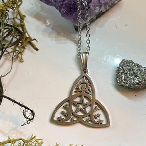 Könnte beinhalten: Eine silberne Halskette mit einem Anhänger, der ein Dreifachmond-Symbol, ein Pentagramm und eine stilisierte menschliche Figur zeigt. Der Anhänger hat die Form eines Triquetra.