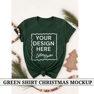 May include: A dark green t-shirt with the text "YOUR DESIGN HERE" in a white frame. The shirt is knotted at the bottom. Decorative elements include a wooden Christmas tree and a string of wooden beads.