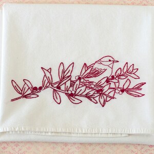 May include: White folded towel with a detailed embroidered design. The design features a bird perched on a branch with leaves and berries, all in a deep red color. The towel is made of a soft, white fabric.