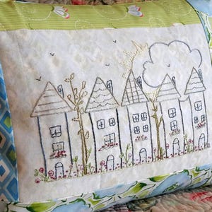 May include: Embroidered cushion featuring a quaint village scene. The design showcases a row of houses with detailed stitching, trees, and flowers. The cushion has a floral and geometric patterned border in blue, green, and white.