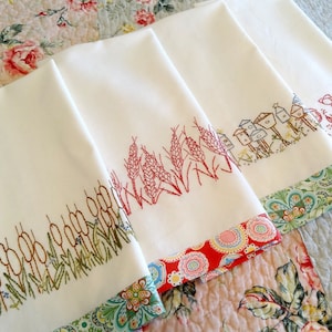 May include: Three white tea towels with embroidered designs. The top towel has a red embroidered design of houses and flowers. The middle towel has a red embroidered design of wheat stalks. The bottom towel has a brown and green embroidered design of tall grass.