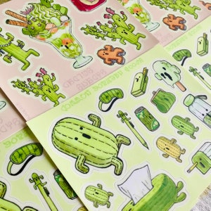 Könnte beinhalten: Eine Bogen mit Aufklebern mit verschiedenen Illustrationen von grünen Kakteen, darunter ein Kaktus mit Schlafmaske, ein Kaktus mit einer Dose Limonade und ein Kaktus mit einem Bleistift. Die Aufkleber befinden sich auf einem hellgrünen Hintergrund mit dem Text "Common Botany Goods" in Schwarz.