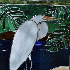 May include: A stained glass window depicting a white egret standing in a blue pond with green leaves. The egret has a long, orange beak and is facing to the right. The leaves are in the background and are partially obscuring the egret.