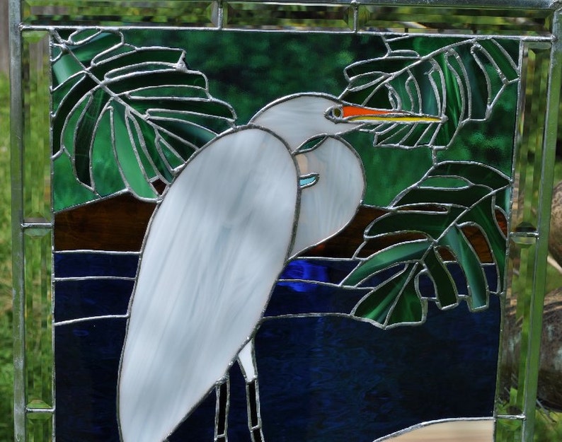 May include: A stained glass window depicting a white egret standing in a blue pond with green leaves. The egret has a long, orange beak and is facing to the right. The leaves are in the background and are partially obscuring the egret.