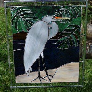 May include: A stained glass window depicting a white egret standing on a sandy beach with green tropical leaves in the background.