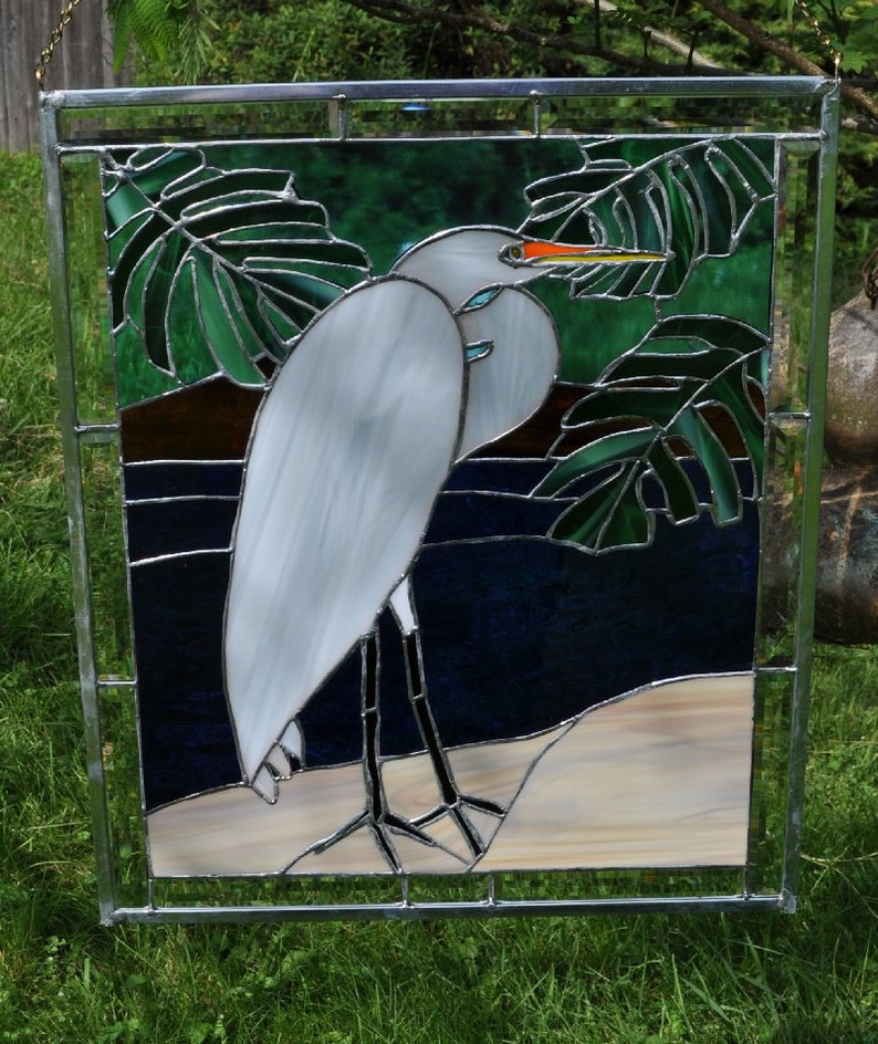 May include: A stained glass window depicting a white egret standing on a sandy beach with green tropical leaves in the background.