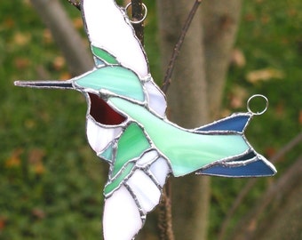 Pode incluir: Um ornamento de beija-flor em vidro colorido com peças de vidro verde, branco e azul. O beija-flor está pendurado em um fio de prata.