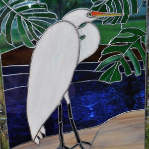 May include: Stained glass window depicting a white egret standing on a sandy beach with blue water and green tropical leaves in the background.