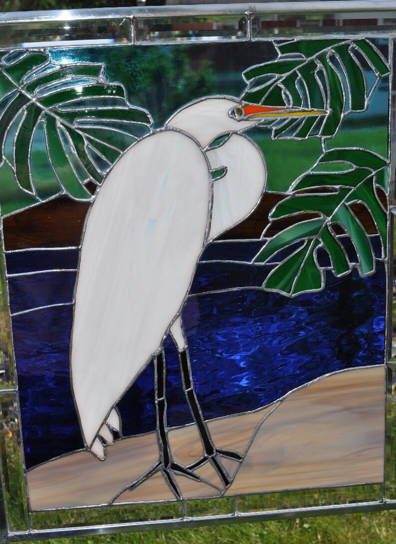 May include: Stained glass window depicting a white egret standing on a sandy beach with blue water and green tropical leaves in the background.