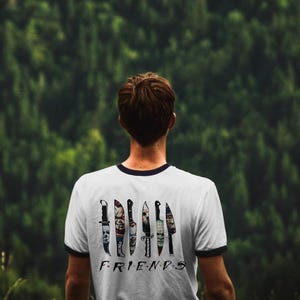 May include: A white t-shirt with a black ringer neckline features a graphic of knives with horror movie characters on them. The text "FRIENDS" is printed below the knives.