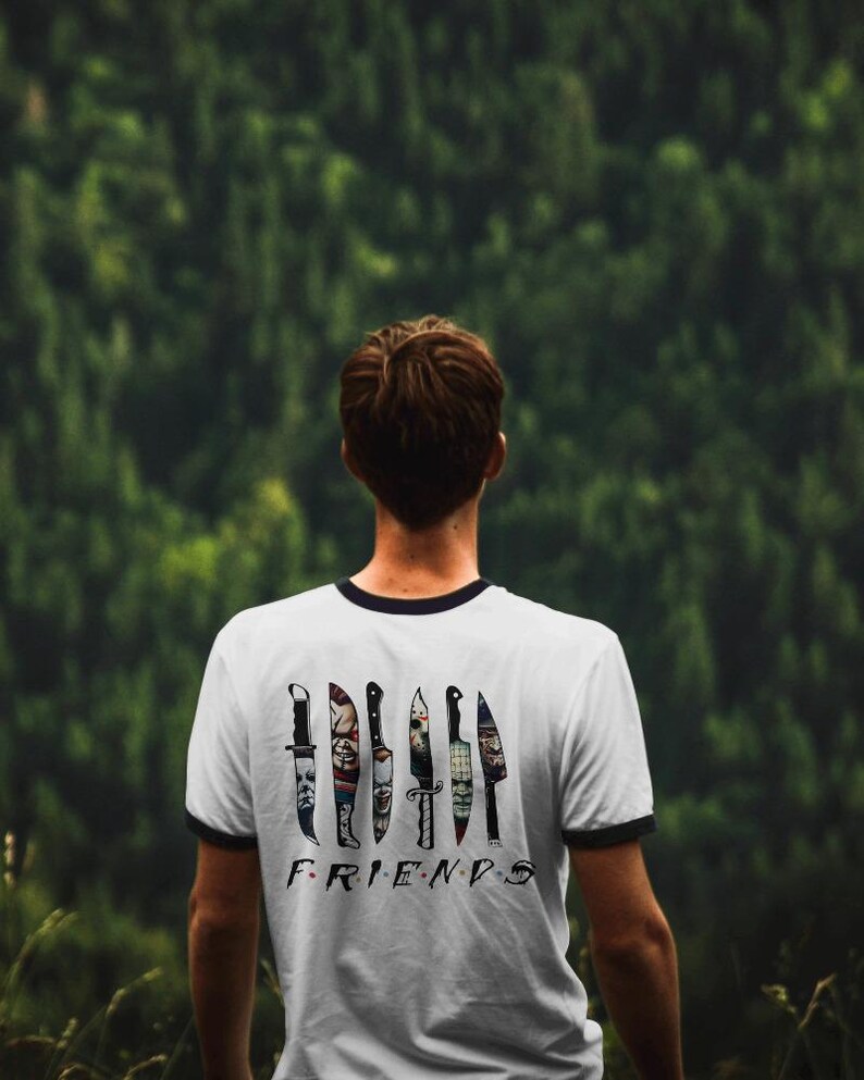 May include: A white t-shirt with a black ringer neckline features a graphic of knives with horror movie characters on them. The text "FRIENDS" is printed below the knives.