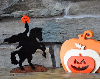 Puede incluir: Decoraciones de Halloween de madera. Una silueta negra del Jinete sin Cabeza sobre un caballo encabritado, sosteniendo una linterna de calabaza. También hay una decoración de calabaza en capas en naranja, blanco y naranja. Las decoraciones están sobre un fondo de piedra.
