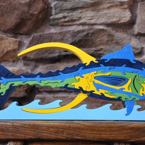 May include: A wooden jigsaw puzzle of a blue, green and yellow fish leaping out of the water. The fish is made up of many interlocking pieces and is a challenging puzzle for adults.