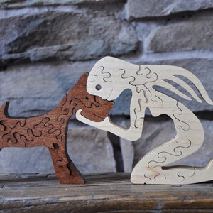 May include: A wooden puzzle of a person kneeling and kissing a dog on the head. The person is made of light wood and the dog is made of dark wood. Both figures are made of interlocking pieces.