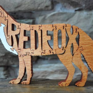 May include: Wooden puzzle of a red fox with the word "RED FOX" cut out of the body. The puzzle is made of light brown wood and has a white painted underbelly.