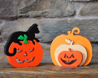 May include: Two wooden pumpkin puzzles, one with a black cat on top, both with carved jack-o'-lantern faces. The pumpkins are orange, with green stems and leaves.