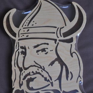 May include: A wooden cutout of a Viking's head with a horned helmet. The cutout is painted in brown and black. The word "VIKINGS" is written below the head.