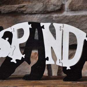 May include: A black and white wooden panda puzzle. The puzzle is made up of the letters P-A-N-D-A, and the pieces fit together to form the shape of a panda.