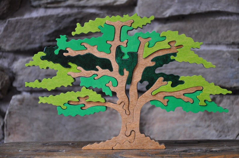 May include: Wooden tree puzzle with a light brown trunk and branches, and green and dark green leaves. The puzzle is made of interlocking pieces, creating a three-dimensional effect. The tree is displayed on a wooden surface.