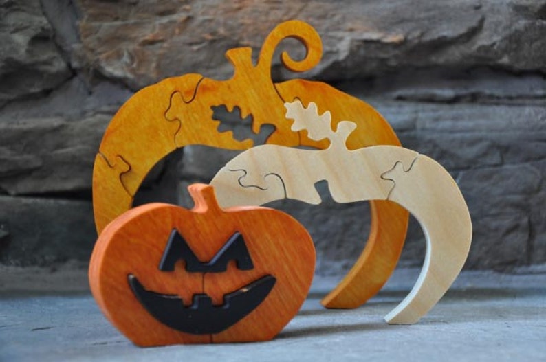 May include: Three wooden pumpkin puzzles, one orange with a jack-o'-lantern face, one orange with a leaf design, and one natural wood with a leaf design.
