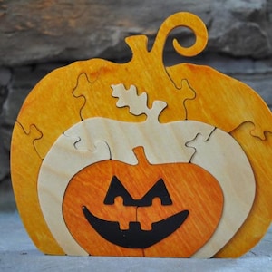 May include: A wooden pumpkin puzzle with a jack-o'-lantern face. The puzzle is made of multiple pieces that fit together to form the pumpkin shape. The pumpkin is painted in shades of orange and yellow.