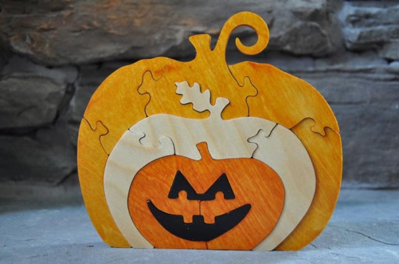 May include: A wooden pumpkin puzzle with a jack-o'-lantern face. The puzzle is made of multiple pieces that fit together to form the pumpkin shape. The pumpkin is painted in shades of orange and yellow.
