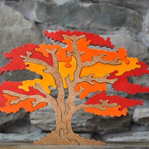 May include: A wooden tree puzzle with a trunk and branches in natural wood tones. The leaves are made of puzzle pieces in shades of red, orange, and yellow. The puzzle is displayed against a gray stone background.