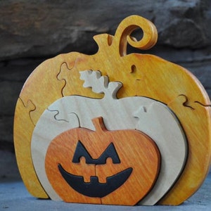 May include: A wooden pumpkin puzzle with three layers. The top layer is yellow, the middle layer is natural wood, and the bottom layer is orange with a black jack-o'-lantern face.