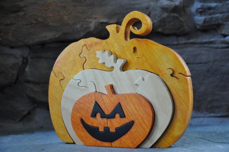 May include: A wooden pumpkin puzzle with three layers. The top layer is yellow, the middle layer is natural wood, and the bottom layer is orange with a black jack-o'-lantern face.