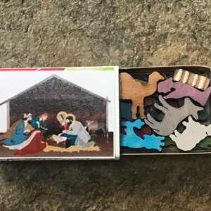 May include: A wooden matchbox with a nativity scene on the front and a set of wooden animal figurines inside. The animals are painted in various colors, including brown, purple, blue, and gray.