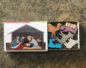 May include: A wooden matchbox with a nativity scene on the front and a set of wooden animal figurines inside. The animals are painted in various colors, including brown, purple, blue, and gray.