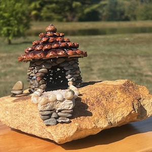 Puede incluir: Una casa de piedra en miniatura con un techo de piña se asienta sobre una gran roca. La casa tiene una pequeña pared de piedra en frente.