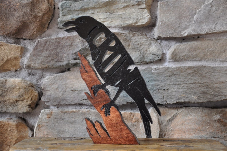 May include: A black wooden crow puzzle with a brown wooden branch. The crow is perched on the branch and has its wings spread out. The puzzle is made of many small pieces that fit together.