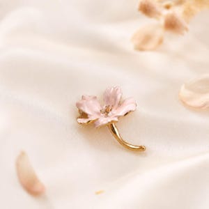 Könnte beinhalten: Eine goldfarbene Metallbrosche in Form einer einzelnen Blume mit fünf hellrosa Emaille-Blütenblättern. Die Mitte der Blume ist mit kleinen goldenen Staubgefäßen versehen und ein geschwungener goldener Stiel ragt aus der Basis hervor.