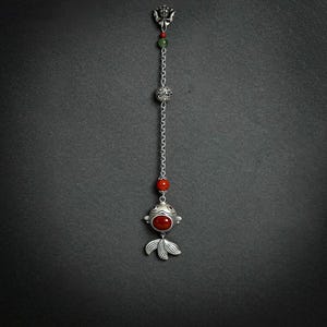 Puede incluir: Un colgante de metal plateado con un diseño de cadena vertical. Incluye un adorno floral, una cuenta verde, una esfera plateada texturizada, una cuenta roja y un adorno en forma de pez con una piedra roja central.