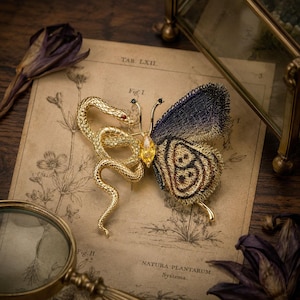 May include: A gold-toned brooch featuring a coiled snake with a red eye on the left side and a butterfly wing on the right side. The butterfly wing is crafted from dark blue and gold metallic thread, with a yellow marquise-cut stone at the center.