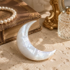 May include: A crescent moon-shaped object with a smooth, iridescent white finish that reflects light. The surface has a pearlescent sheen, displaying subtle variations in tone across its curved form, creating a luminous appearance.