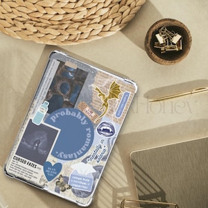 Puede incluir: Una funda transparente para tableta decorada con pegatinas y texto. Las pegatinas incluyen un drag&oacute;n dorado, una tirita que dice "Star Crossed" y un coraz&oacute;n azul. El texto incluye "Probably Romantasy" y "Cursed Fates".