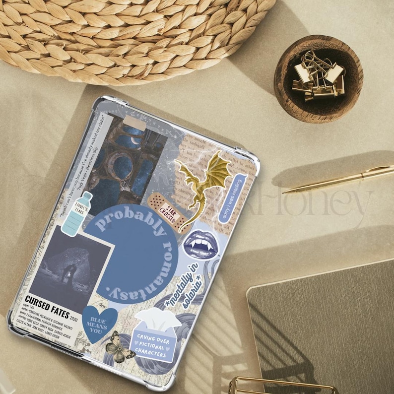 Puede incluir: Una funda transparente para tableta decorada con pegatinas y texto. Las pegatinas incluyen un drag&oacute;n dorado, una tirita que dice "Star Crossed" y un coraz&oacute;n azul. El texto incluye "Probably Romantasy" y "Cursed Fates".