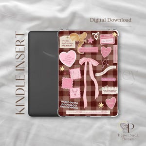 May include: A Kindle e-reader featuring a decorative insert with a dark red and brown plaid background. The design includes a central pink ribbon, a gold dragon illustration, various pink heart-shaped stickers, and text snippets including "Probably Reading Romantasy" and "Finders Keepers" scattered across the surface.