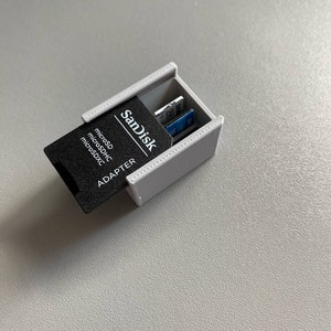 Peut inclure: Un support de carte SD gris imprimé en 3D avec un adaptateur SanDisk noir et plusieurs cartes SD à l'intérieur. L'adaptateur porte l'inscription "microSD, microSDHC, microSDXC ADAPTER". Le support est ouvert, révélant les cartes.