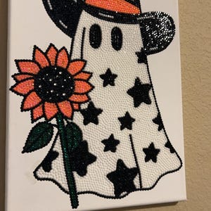 May include: A diamond art canvas featuring a white ghost wearing a black cowboy hat with an orange band. The ghost is decorated with black stars and holds a large orange sunflower with a green stem and leaves, all created with sparkling beads.