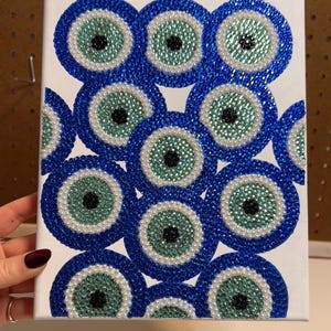 May include: A white canvas featuring a pattern of blue, green, and white beaded evil eye designs. The eyes have a blue outer ring, a white inner ring, a green center, and a black pupil. The artwork is held by a hand.