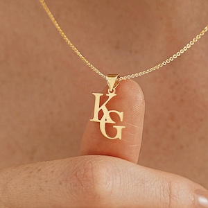 以下が含まれることがあります： 「KG」の文字が絡み合ったペンダントが付いたゴールドカラーのネックレス。繊細なチェーンとペンダントがニュートラルな背景に映え、ジュエリーのデザインを際立たせています。このネックレスはパーソナライズされたアクセサリーです。